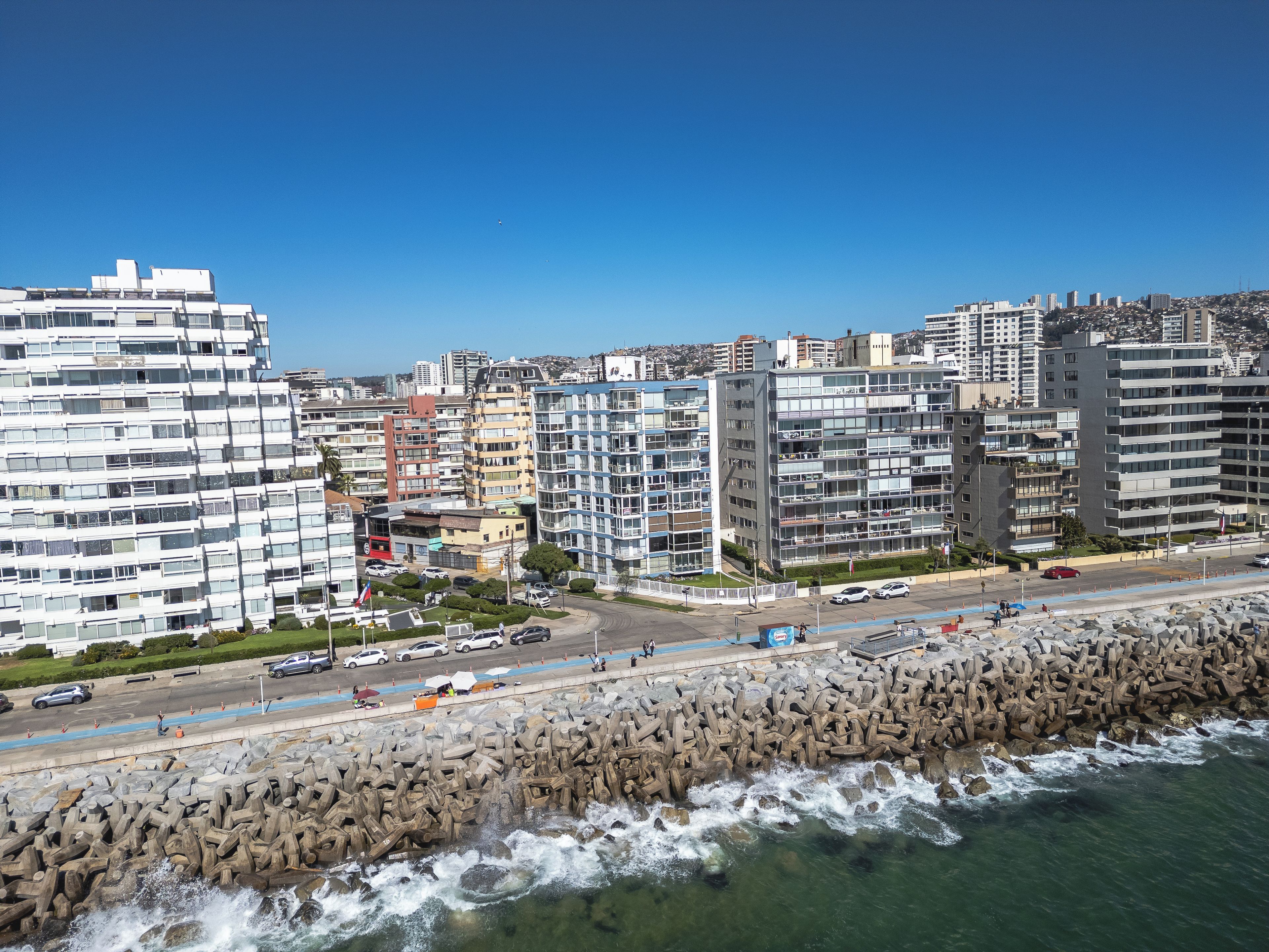 A coastal cityscape with modern buildings, a rocky breakwater, and a clear blue sky.