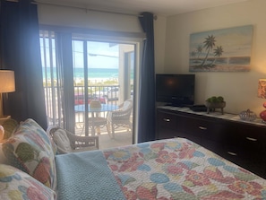 A hotel room with a bed, a television, a desk with a computer, a window with a view of the beach, and a balcony with a table and chairs. - Ocean View! Come Escape with Us! (Bradenton Beach)