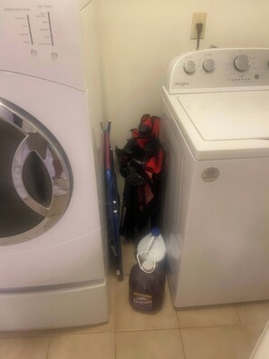 Un área de lavandería con lavadora de carga frontal y secadora. Hay una botella de detergente y un bulto rojo y negro en el piso.