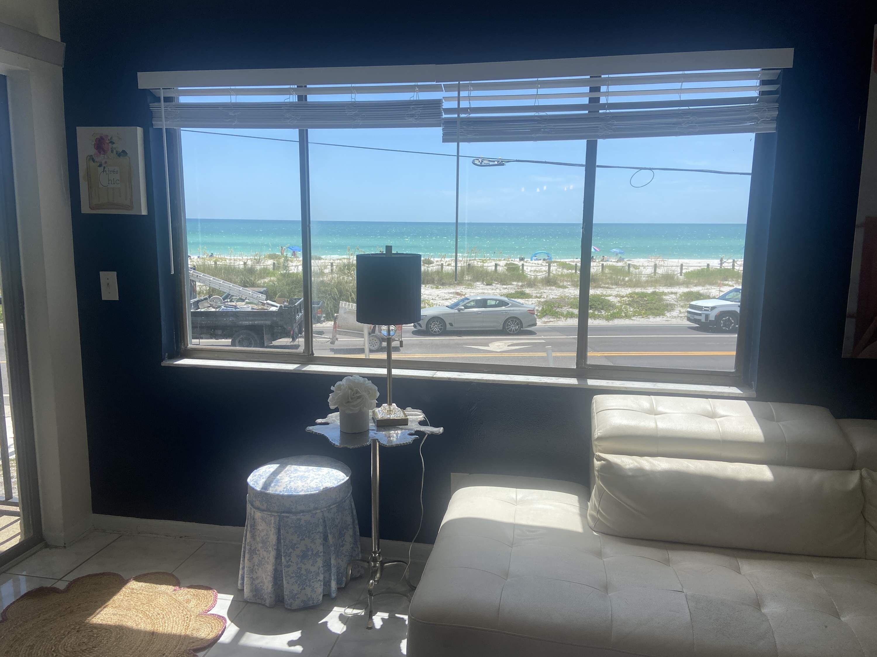 Ein Zimmer mit großem Fenster mit Blick auf einen Strand, ein weißes Sofa, ein kleiner runder Tisch und eine Lampe.