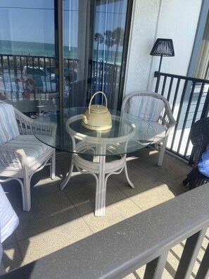 Un tavolo con piano in vetro, una teiera color crema, circondato da sedie in vimini, su un balcone con vista sul mare.