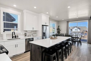 A modern kitchen with white cabinetry, a central island with bar stools, and a dining area with a view of the outside. - Lakeview Gamehouse | Ideal Beach Access+EV Charger (Garden City)