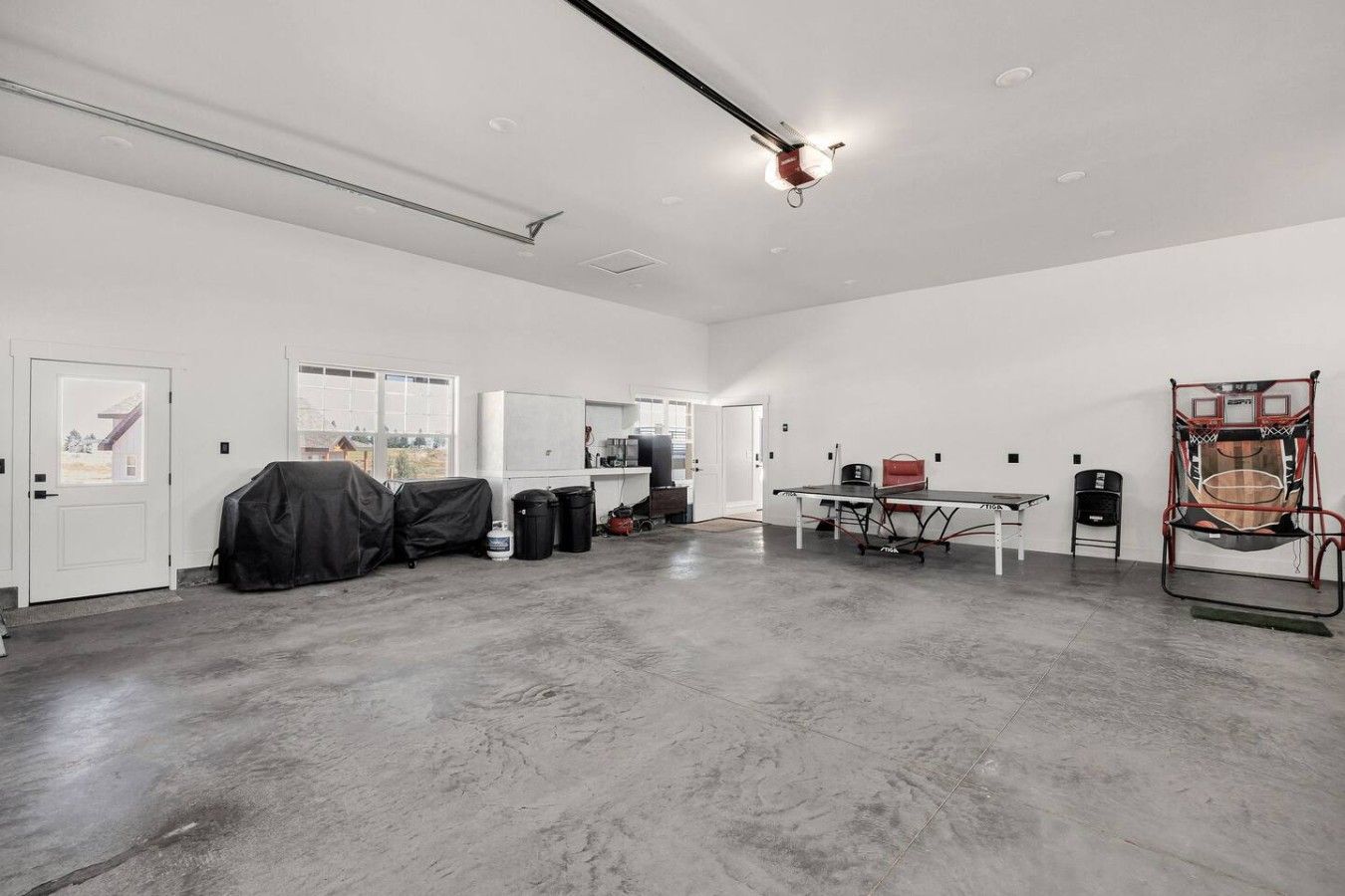 A spacious room with a concrete floor, a table with chairs, a basketball hoop, and a door with a view.