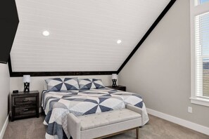 A bedroom with a sloped ceiling, a bed with a geometric patterned bedspread, two bedside tables with lamps, and a window with blinds. - Lakeview Gamehouse | Ideal Beach Access+EV Charger (Garden City)