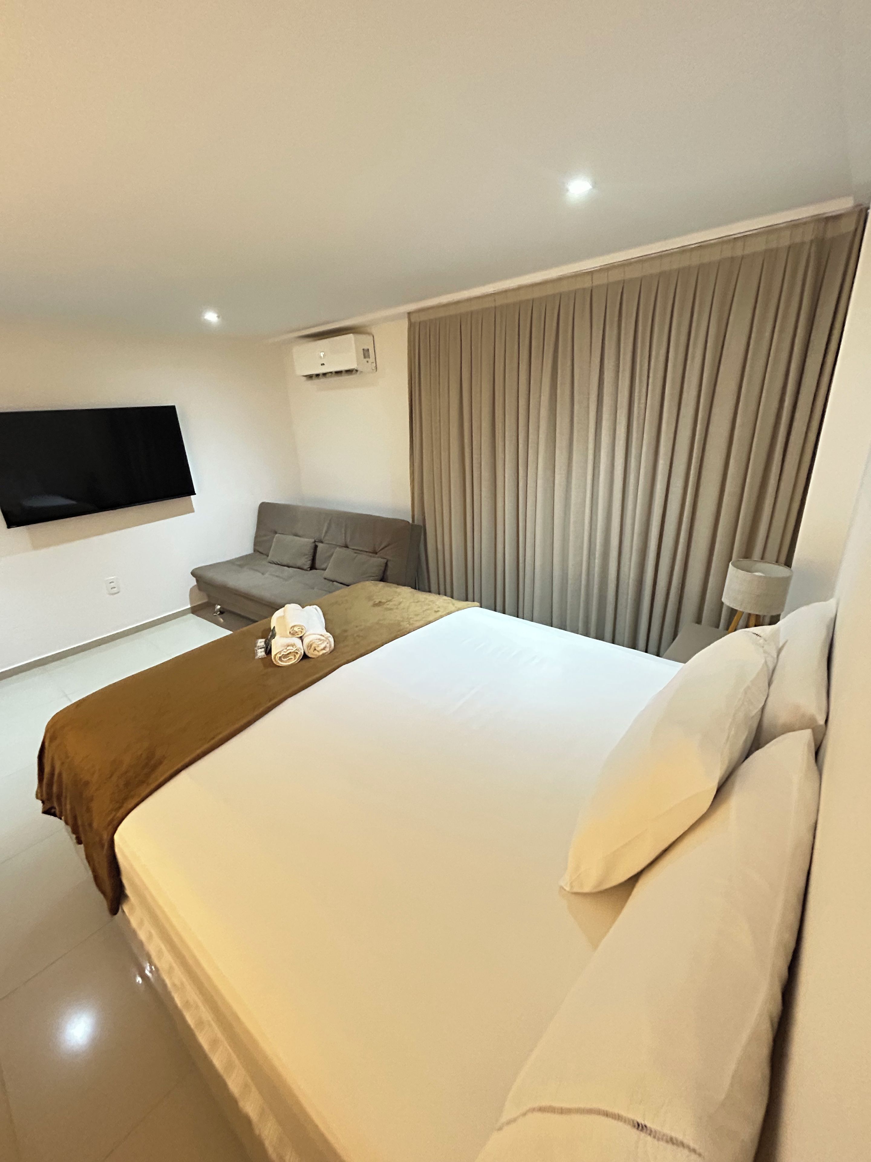 A modern hotel room with a large bed, a sofa, a flat-screen TV, and an air conditioning unit.