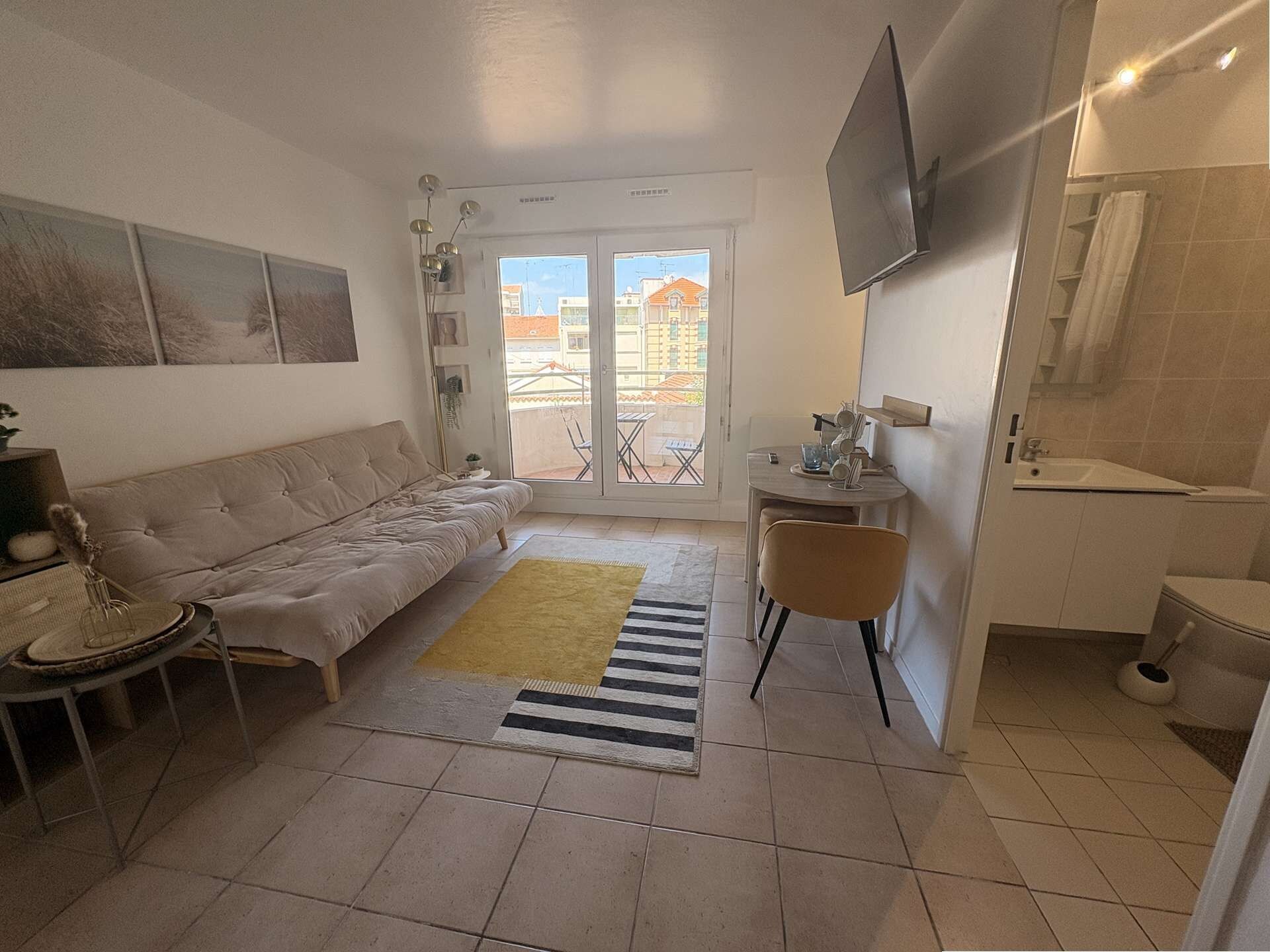 A compact living space with a sofa, a small table, a chair, a TV mounted on the wall, and a balcony with a view of buildings.