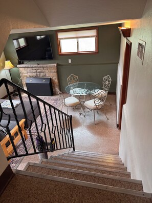 Un escalier mène à un espace de vie comprenant une cheminée, une table à manger avec des chaises et un téléviseur fixé au mur.