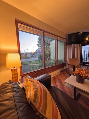 Ein Wohnzimmer mit großem Fenster, einer Couch, einer Lampe und einem Tisch mit Blumenvase.