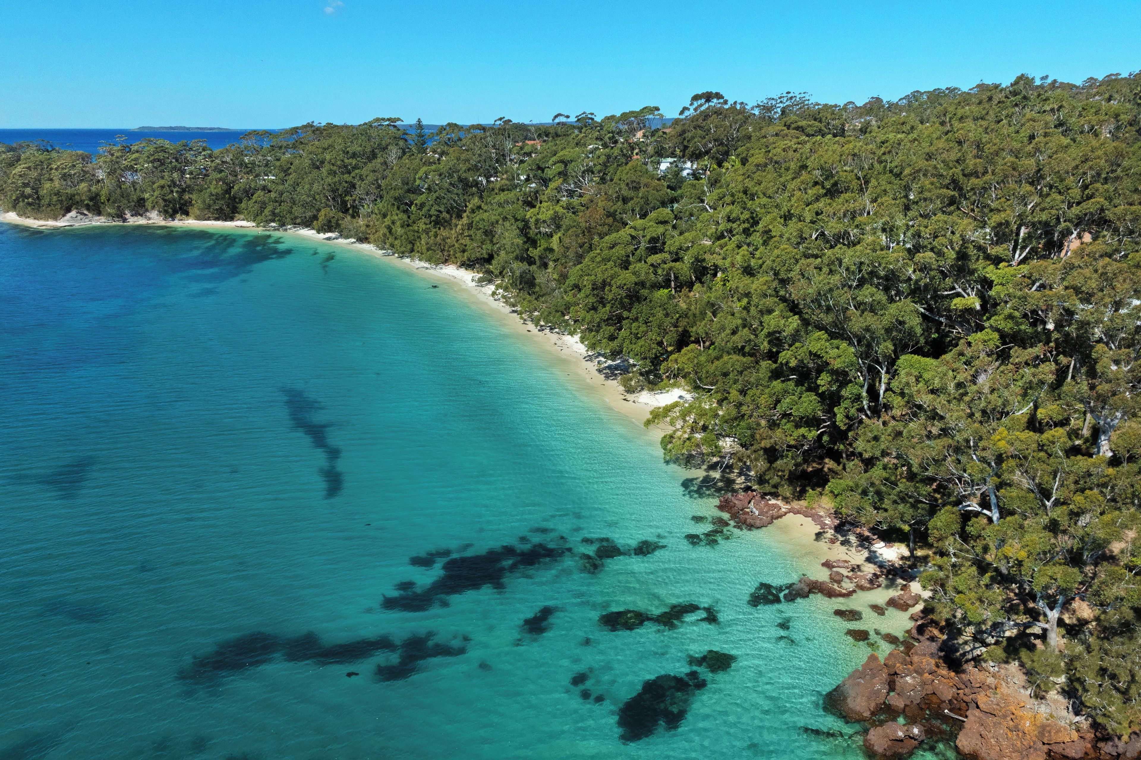 An aerial view of a coastal area with a clear turquoise sea, a sandy beach, and dense green forest.