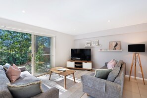Ein modernes Wohnzimmer mit einem Flachbildfernseher, grauen Sofas, einem Couchtisch und einer großen Schiebeglastür, die zu einem Garten führt.