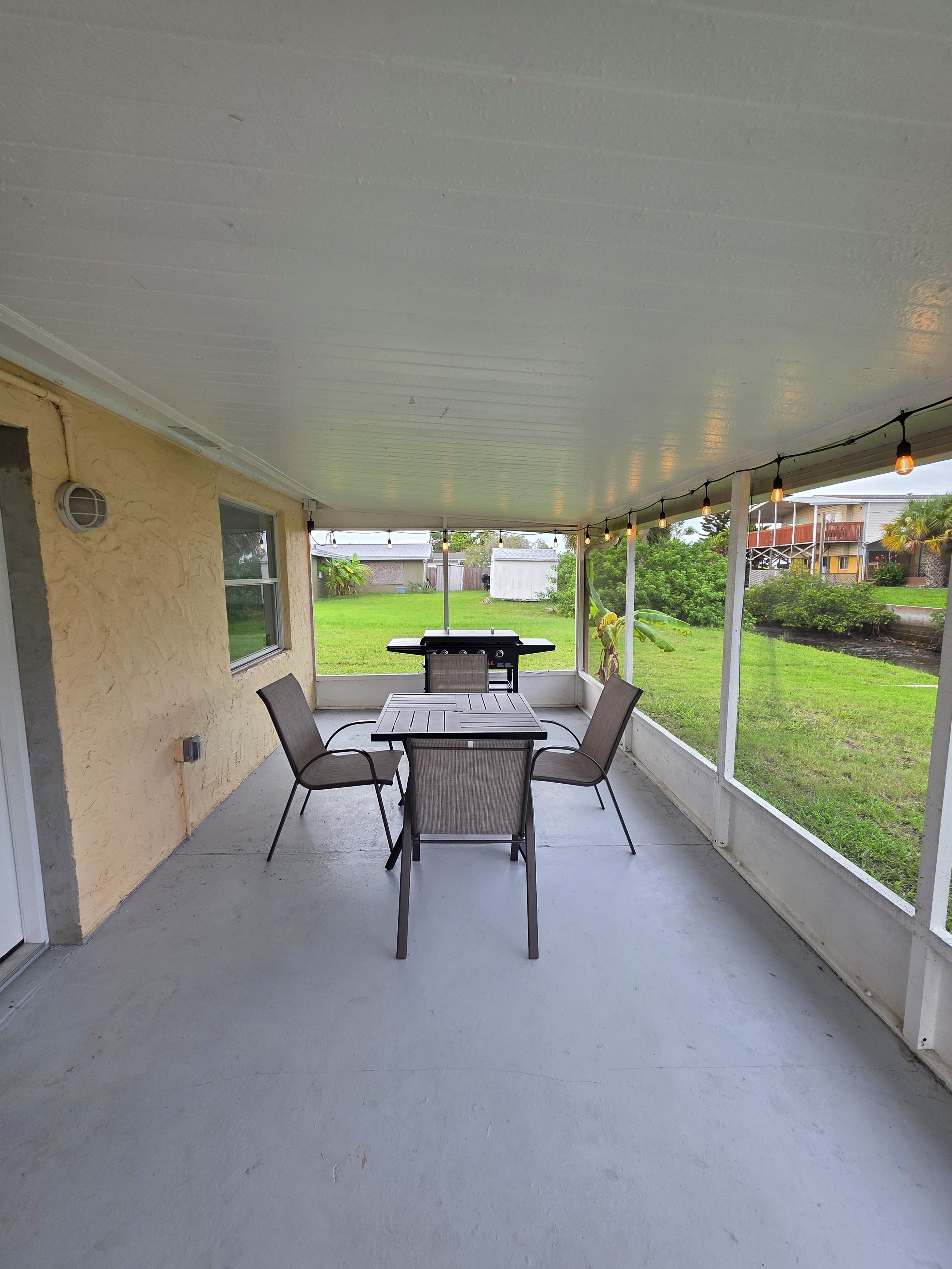 Een overdekt terras met een tafel, stoelen, een barbecue en uitzicht op de achtertuin.