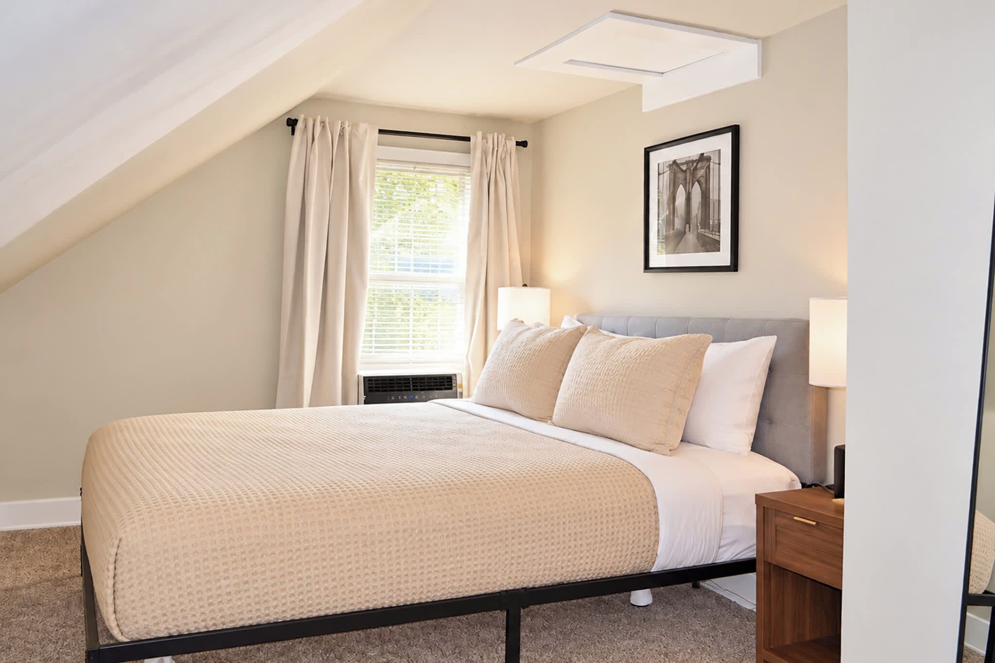 A bedroom with a bed, a nightstand, a window with curtains, a wall-mounted picture, and a sloped ceiling.