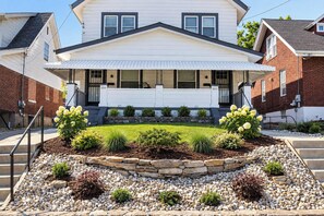 A two-story house with a front porch, a stone retaining wall, and a landscaped garden with shrubs and a small tree. - Cincy 5BR Retreat | Xavier + Zoo | Sleeps 7 (Cincinnati)