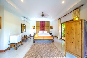Quarto com cama, mesas de cabeceira, guarda-roupa, ventilador de teto e vista para o verde lá fora.