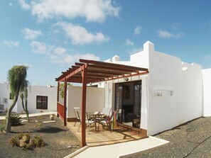 Ein überdachter Außenbereich mit Tisch und Stühlen, einer Pergola und einem weißen Gebäude mit einer Tür.