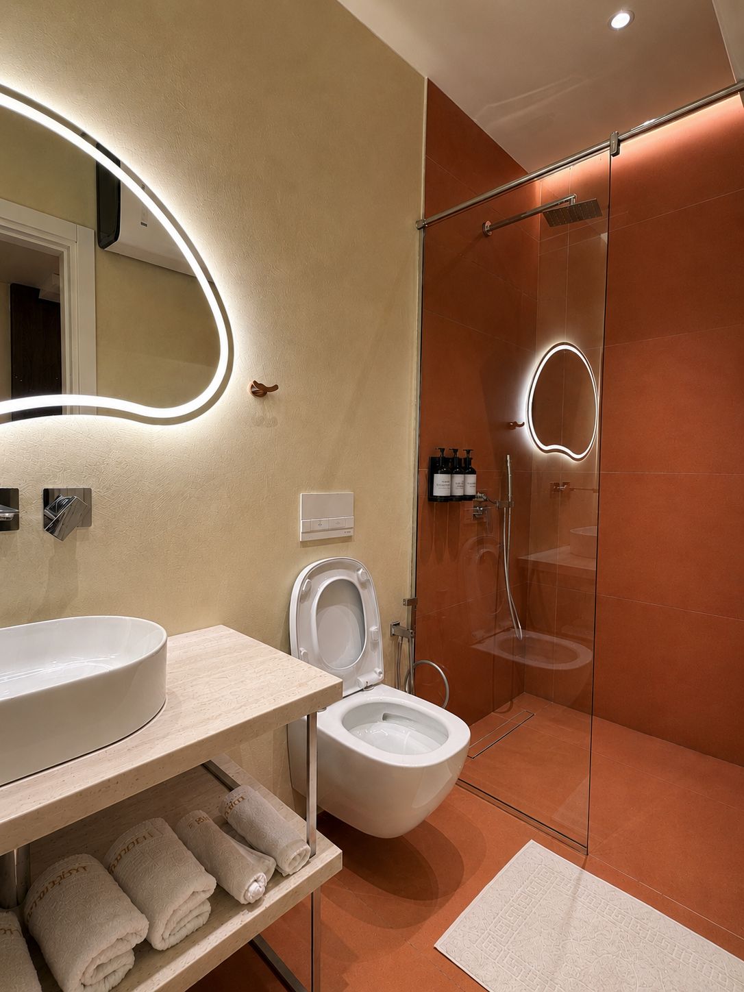 Un baño moderno con un espejo grande, inodoro, ducha y tocador con lavamanos y toallero.