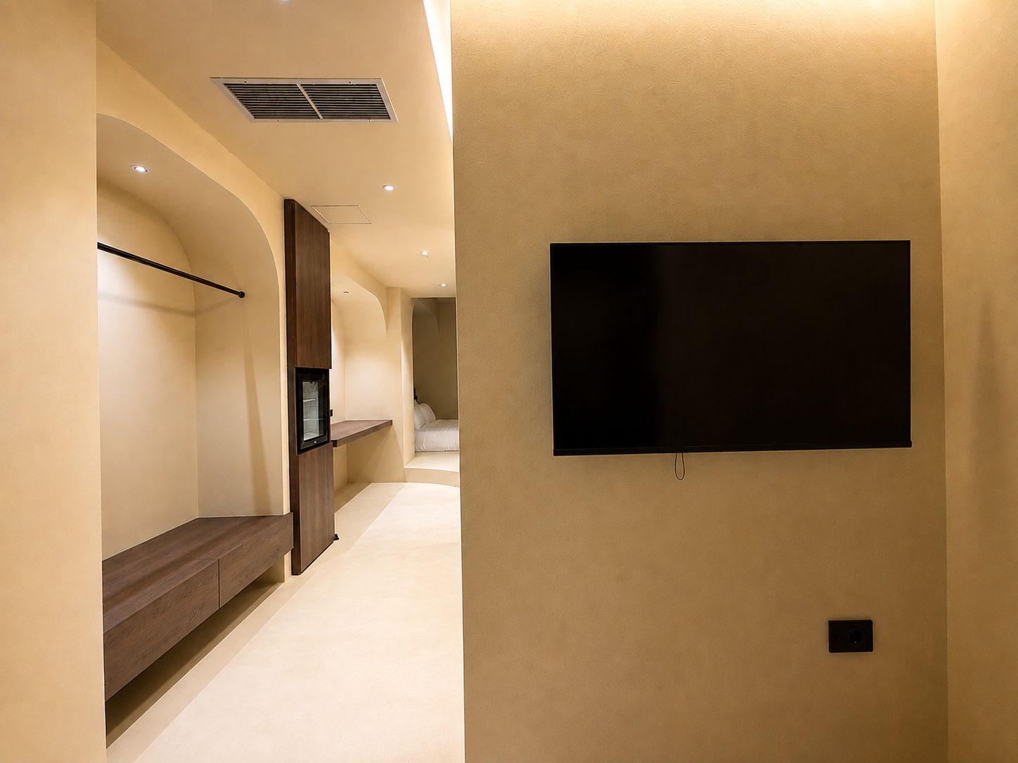 A hallway with a flat-screen TV mounted on the wall, a wooden bench, and a recessed ceiling light.