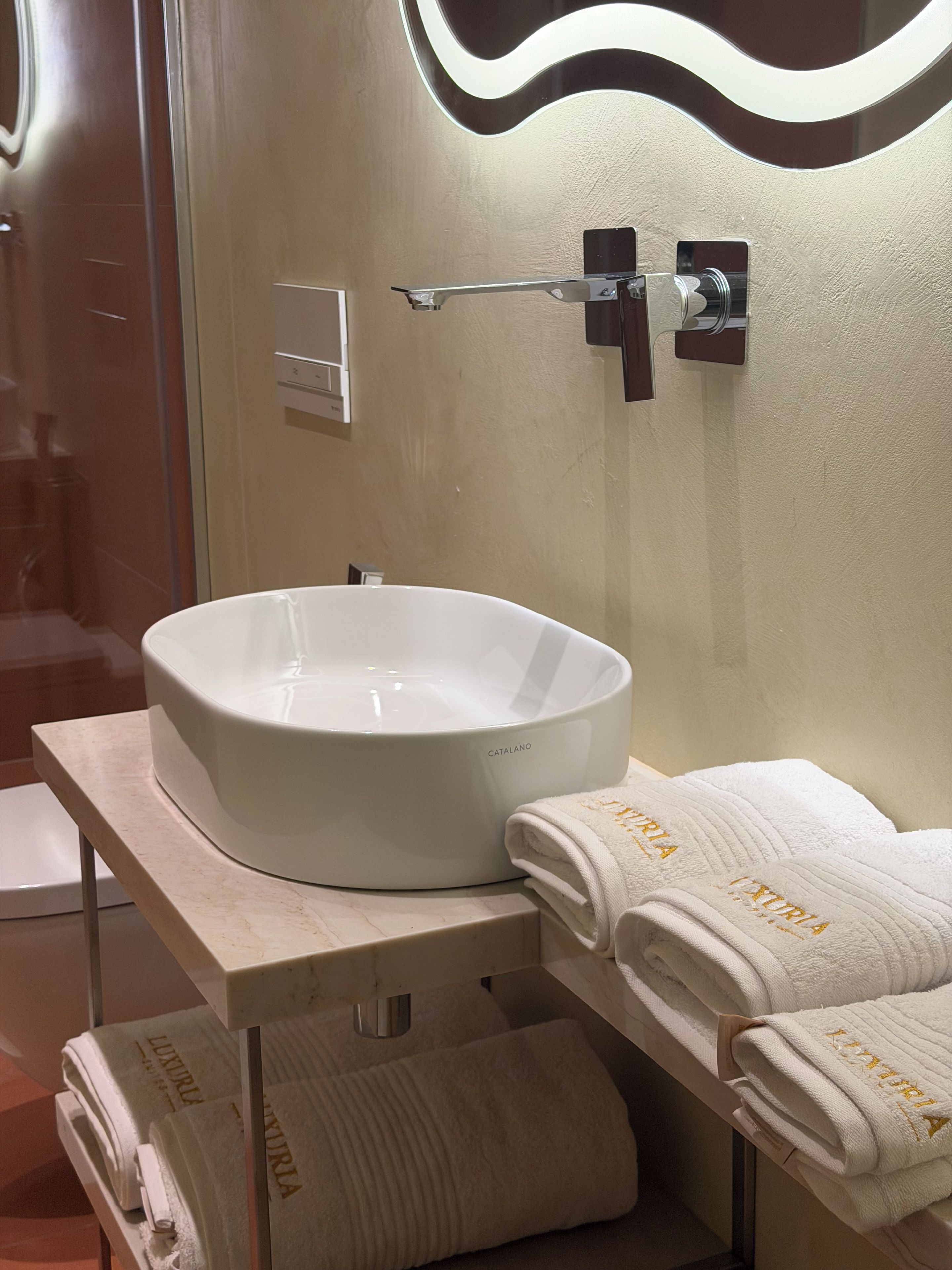 A bathroom with a white vessel sink, a towel rack with folded towels, and a mirror with a wavy design.