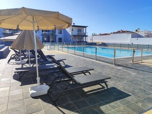 Un espace aménagé au bord de la piscine, avec des chaises longues et des parasols, une piscine, et un bâtiment en arrière-plan.