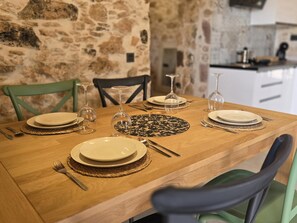 A wooden dining table set with plates, cutlery, and wine glasses, surrounded by chairs in a stone-walled room. - La Montañeta - Tefia Suites (Tefía)