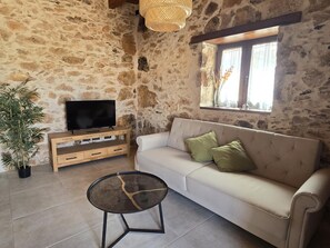A stone-walled room with a sofa, a television on a wooden stand, a small table, and a window with curtains. - La Montañeta - Tefia Suites (Tefía)
