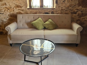 A beige sofa with two green cushions, a glass coffee table with a reflective surface, and a stone wall with a window above. - La Montañeta - Tefia Suites (Tefía)