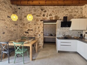 A rustic interior with a stone wall, wooden ceiling, a dining table with chairs, a kitchen with white cabinets, and pendant lights. - La Montañeta - Tefia Suites (Tefía)