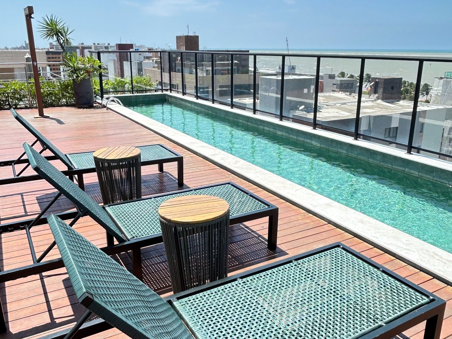 Piscina no terraço com espreguiçadeiras e mesas, delimitada por guarda-corpo de vidro.