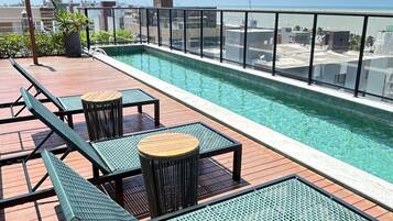 Piscina no terraço com espreguiçadeiras e mesas, delimitada por guarda-corpo de vidro.