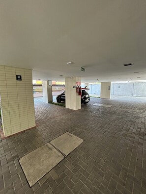 Uma área de estacionamento coberta com piso de azulejos, um extintor de incêndio fixado em um pilar, e um carro estacionado ao fundo.