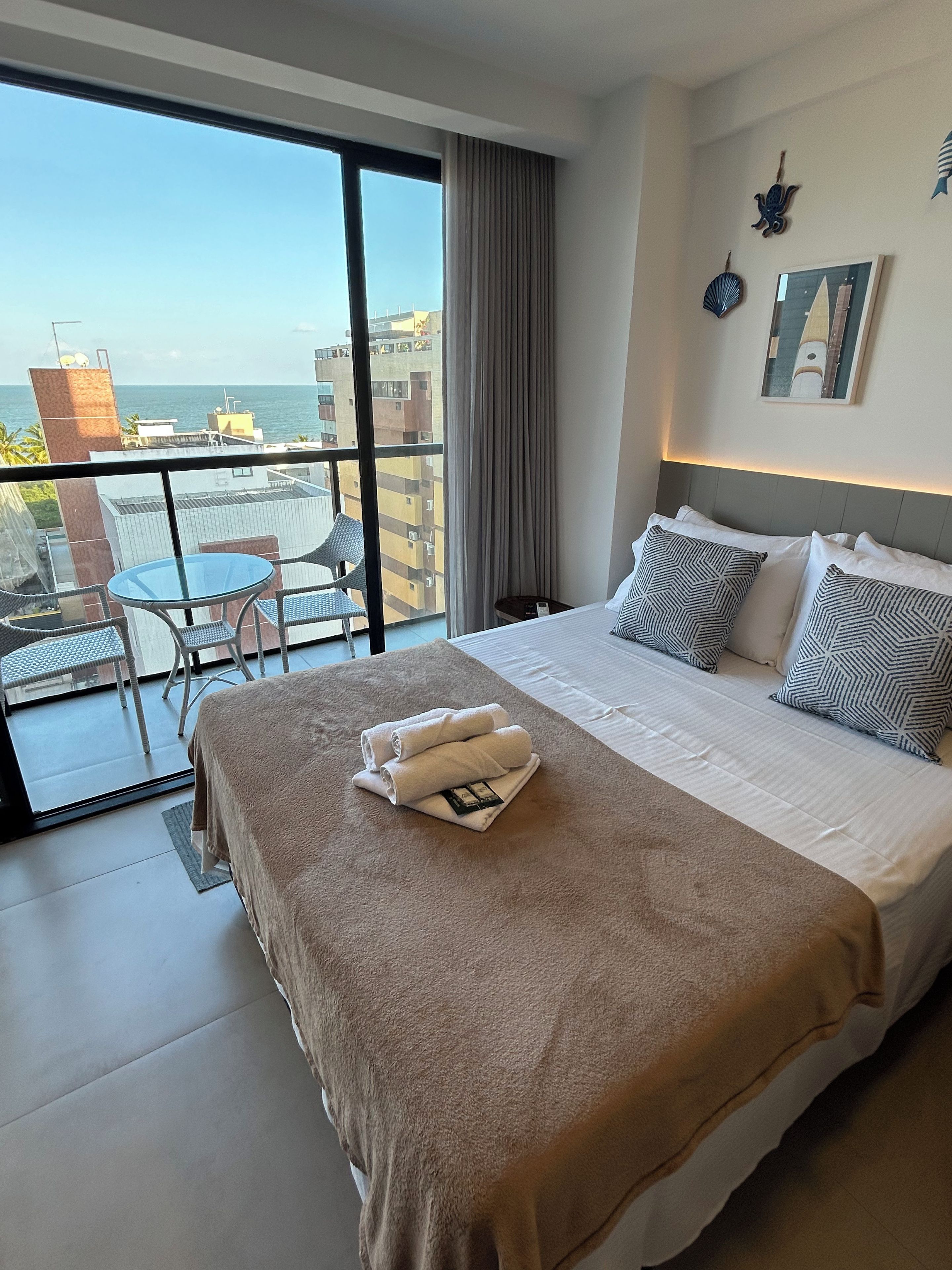 Quarto de hotel com uma cama grande, varanda com mesa e cadeiras, e vista para o mar.