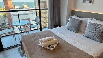 Quarto de hotel com uma cama grande, varanda com mesa e cadeiras, e vista para o mar.
