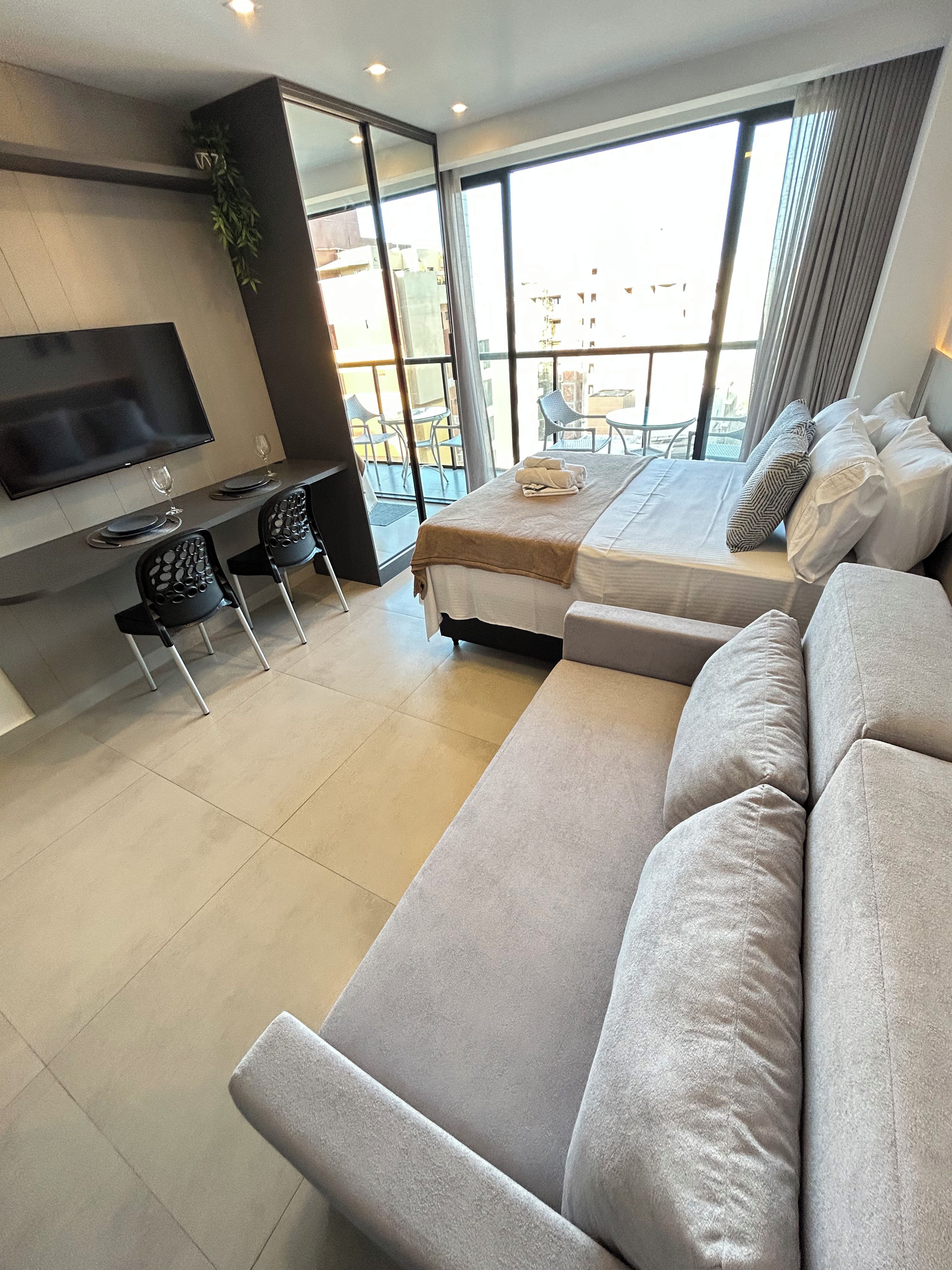 Uma sala de estar moderna com sofá, televisão de tela plana, área de jantar com cadeiras e mesa, e varanda com vista.
