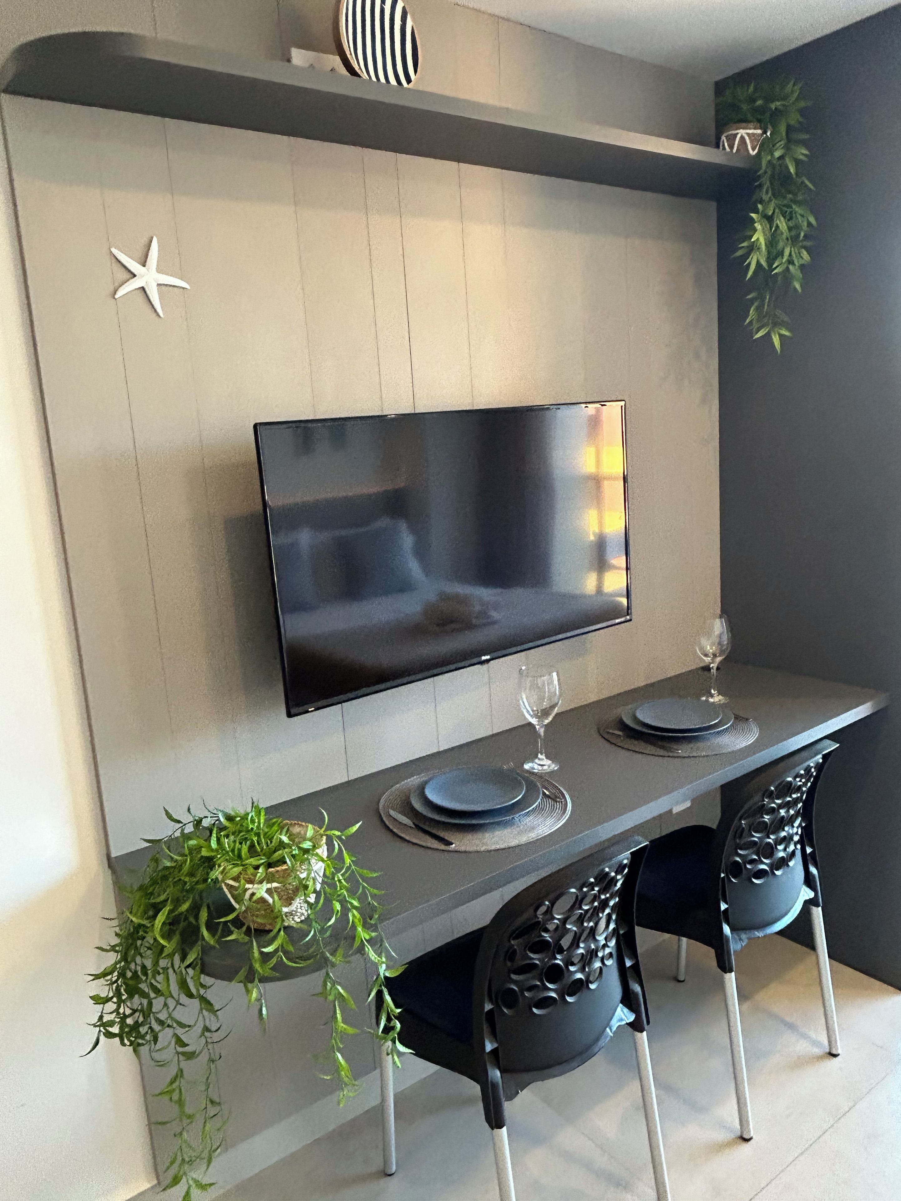 Uma sala de jantar moderna com televisão de tela plana na parede, uma mesa de jantar comprida com duas cadeiras e elementos decorativos como uma estrela e uma planta.