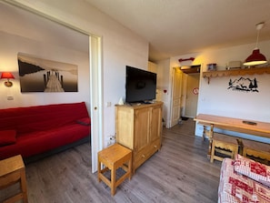 A cozy living room with a red sofa, a television on a wooden cabinet, a small wooden table, and a wall-mounted shelf with a lamp. - Appartement 6 pers avec balcon et vue sur le Queyras – Résidence Betelgeuse (Risoul)
