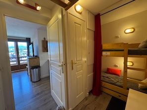 A room with a bunk bed, a door with a red curtain, and a view of a balcony. - Appartement 6 pers avec balcon et vue sur le Queyras – Résidence Betelgeuse (Risoul)