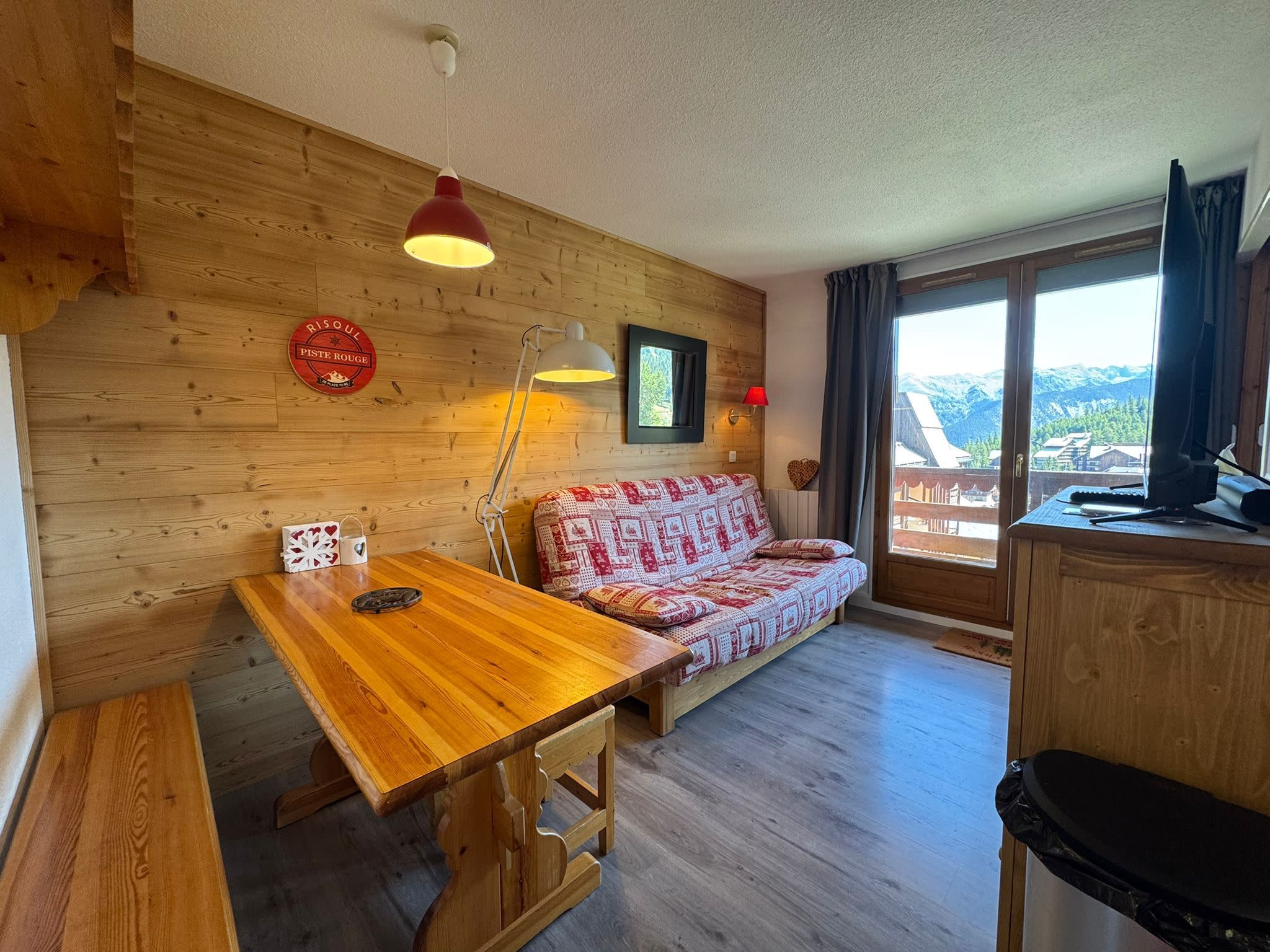 A cozy room with a wooden wall, a sofa, a dining table, and a view of mountains through a window.