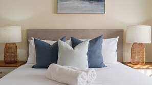 A neatly made bed with a white duvet, multiple pillows in shades of blue and white, and a rolled white towel. There are two wicker lamps on either side of the bed.