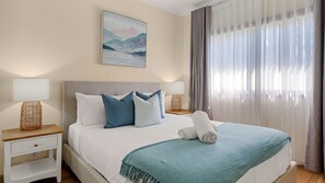 A hotel room with a bed, two bedside tables, a lamp, a painting, and a window with curtains. - The Lazy Pelican with Fireplace, Deck & Lake Views (Wyongah)