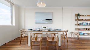 A dining area with a wooden table set for four, surrounded by wicker chairs, a white ceiling light, and a coastal landscape painting above. - The Birdsnest Bondi (Waverley)