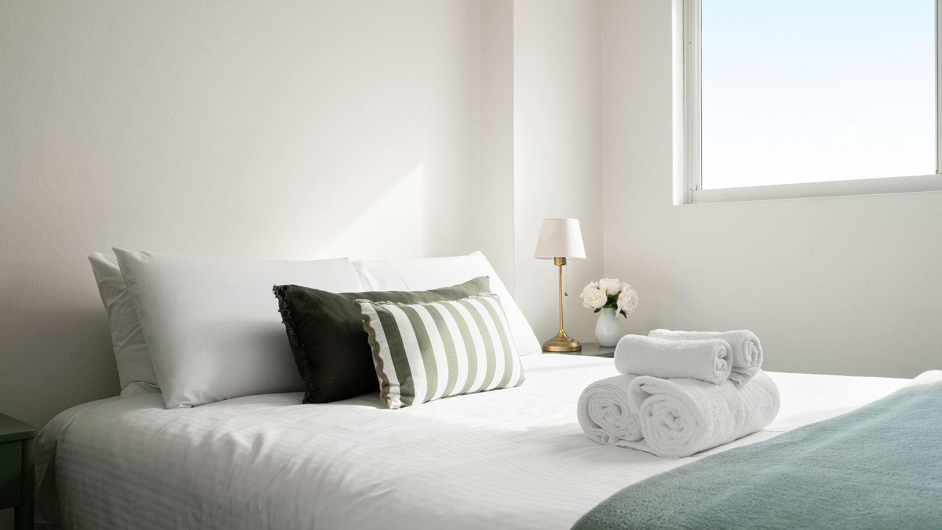 Un lit bien fait, avec une literie blanche, un oreiller à rayures vertes et blanches, et des serviettes blanches pliées.