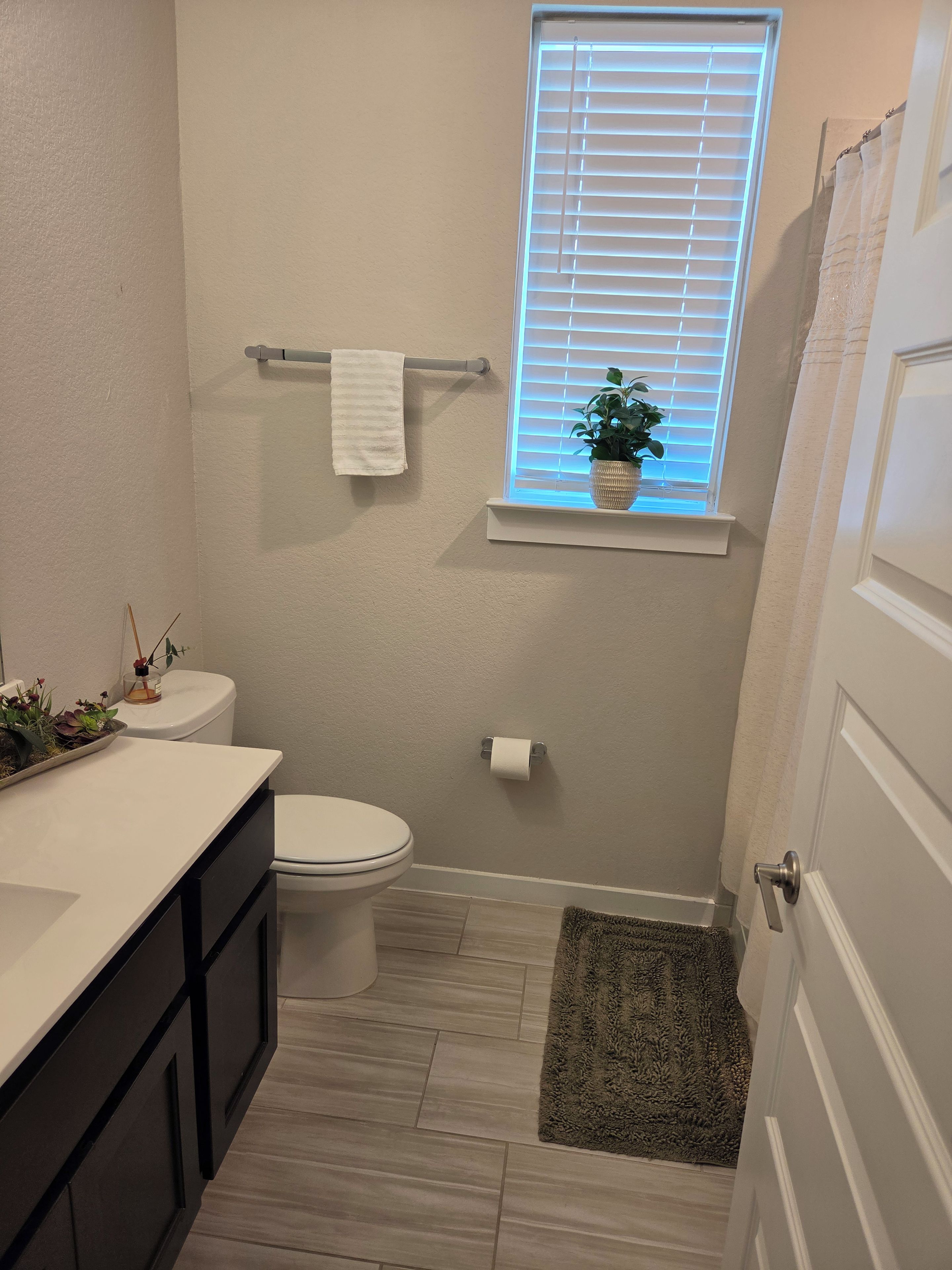 A bathroom with a toilet, a sink with a countertop, a window with blinds, a towel rack, a potted plant, and a shower curtain.