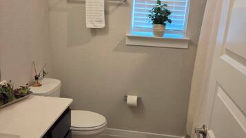 A bathroom with a toilet, a sink with a countertop, a window with blinds, a towel rack, a potted plant, and a shower curtain.