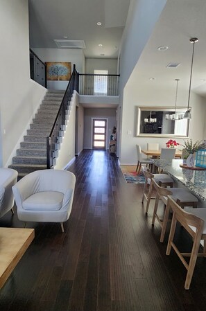 Un interior espacioso con una escalera que conduce a un segundo piso, una zona de comedor con taburetes de bar y una cocina con un amplio ventanal.