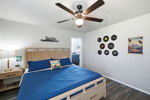 Une chambre à coucher comprenant un lit, un ventilateur de plafond, une table de chevet, une lampe, un mur décoré d’un motif représentant une vue panoramique de la ville, et un autre mur orné de disques vinyle.