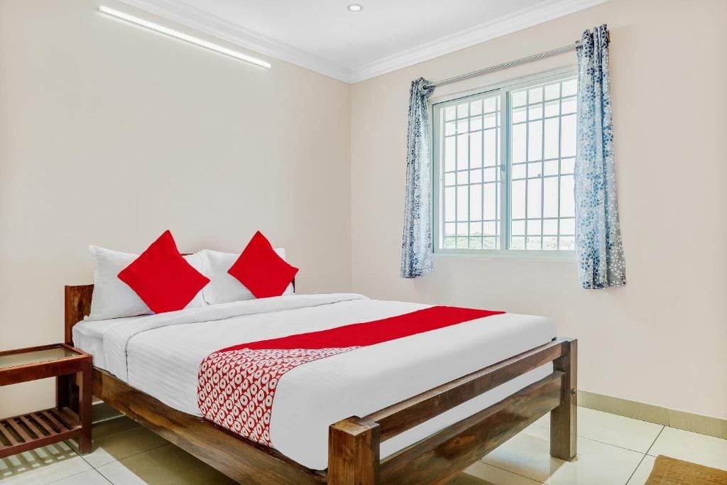 A hotel room with a wooden bed, white bedding, red pillows, a window with blue curtains, and a small wooden nightstand.