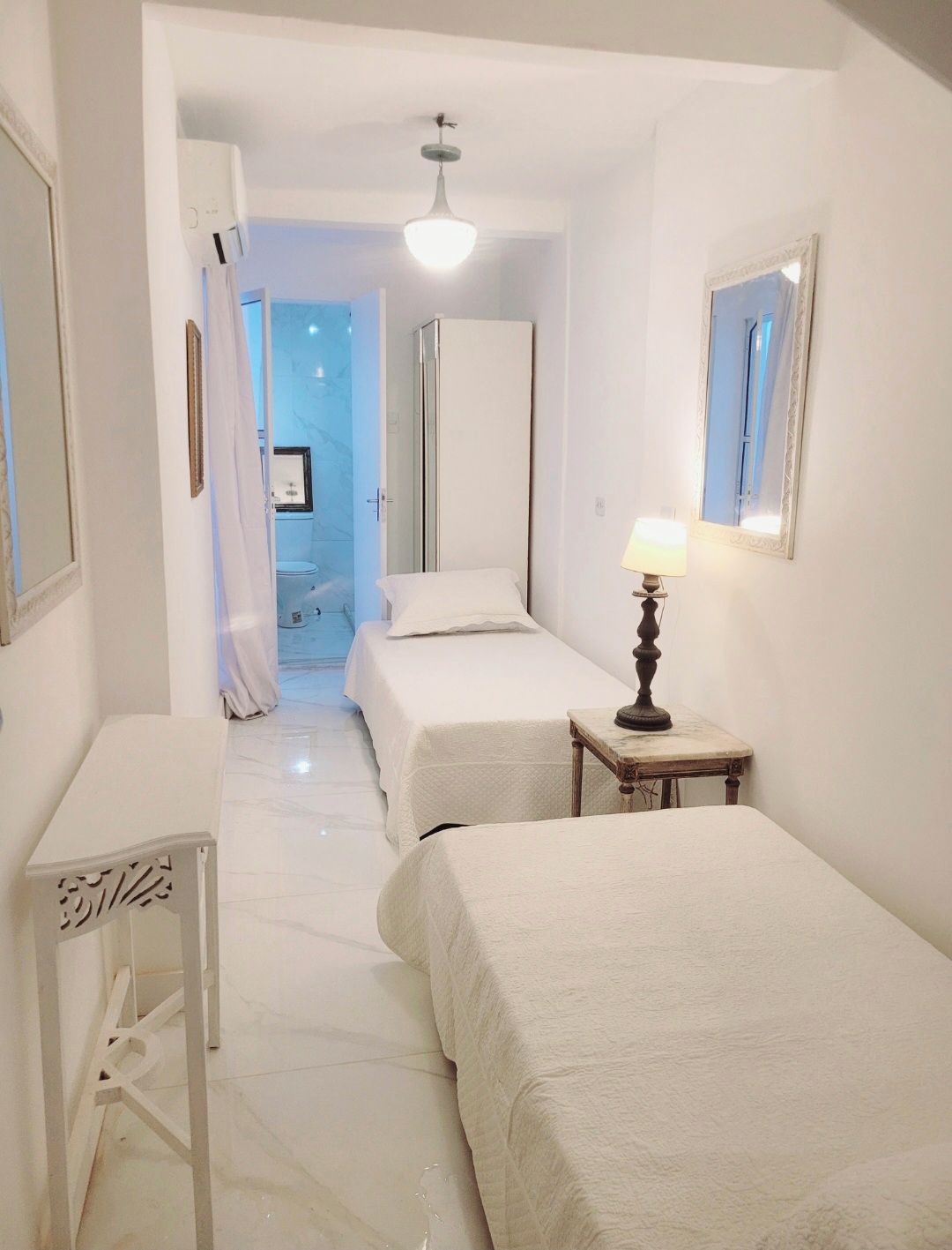 A hotel room with two beds, a bedside table with a lamp, a wardrobe, and a bathroom visible through an open door.