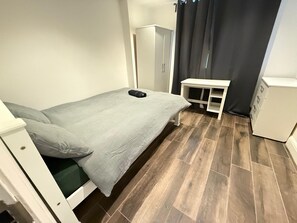 A small, single-bed room with a grey bedspread, a white bedside table, a white wardrobe, and a dark curtain. - Entire Luxury 2 Bedroom Apartment, Free Parking. (South Croydon)