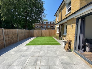 A backyard with a paved patio, a green lawn, a wooden fence, and a residential building in the background. - Entire Luxury 2 Bedroom Apartment, Free Parking. (South Croydon)
