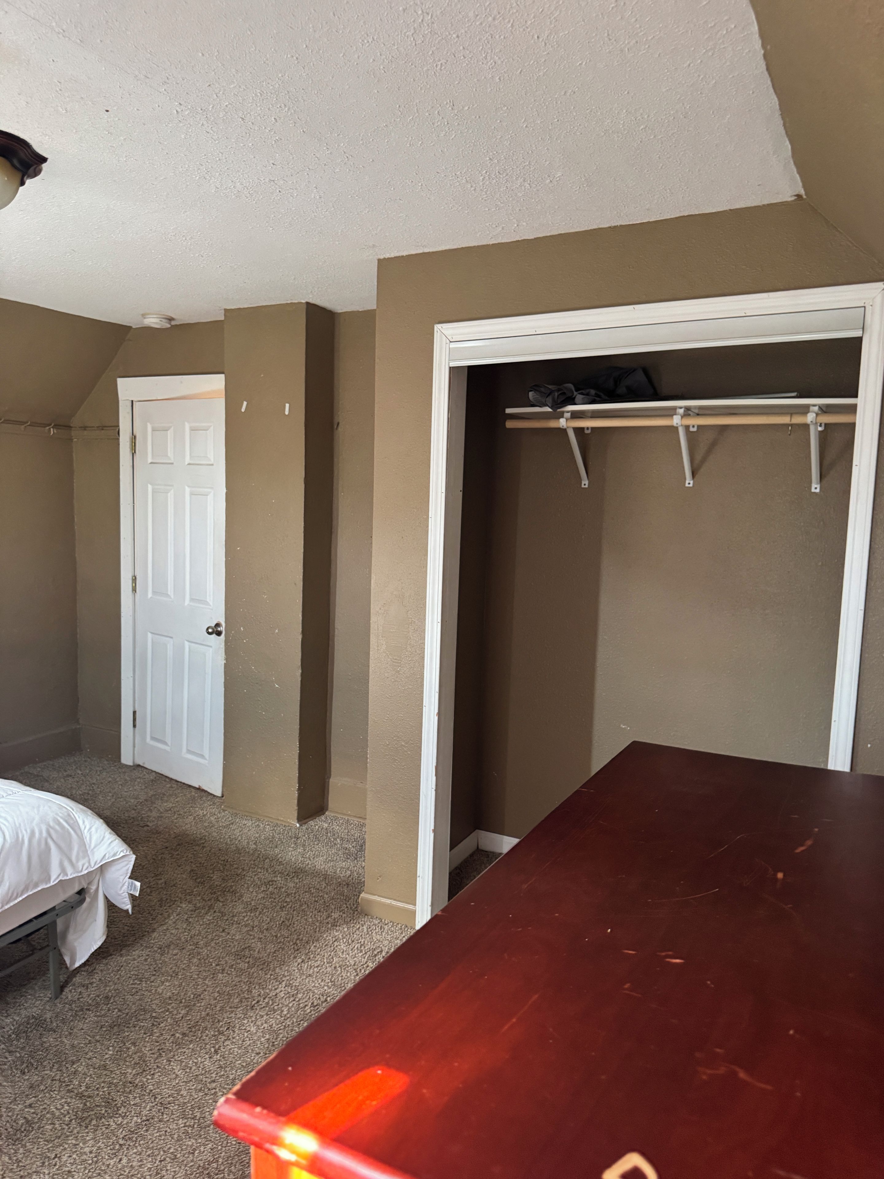 A bedroom with a bed, a wooden dresser, a closet with a rod, and a door.